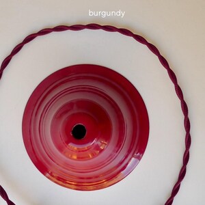 May include: A glossy burgundy metal ceiling canopy with a central circular opening, accompanied by a matching burgundy twisted fabric electrical cord arranged in a large circle around the canopy. The text burgundy is visible above.