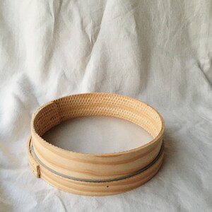 May include: A round wooden flour sifter with a gray mesh lining. The sifter is sitting on a white fabric surface.