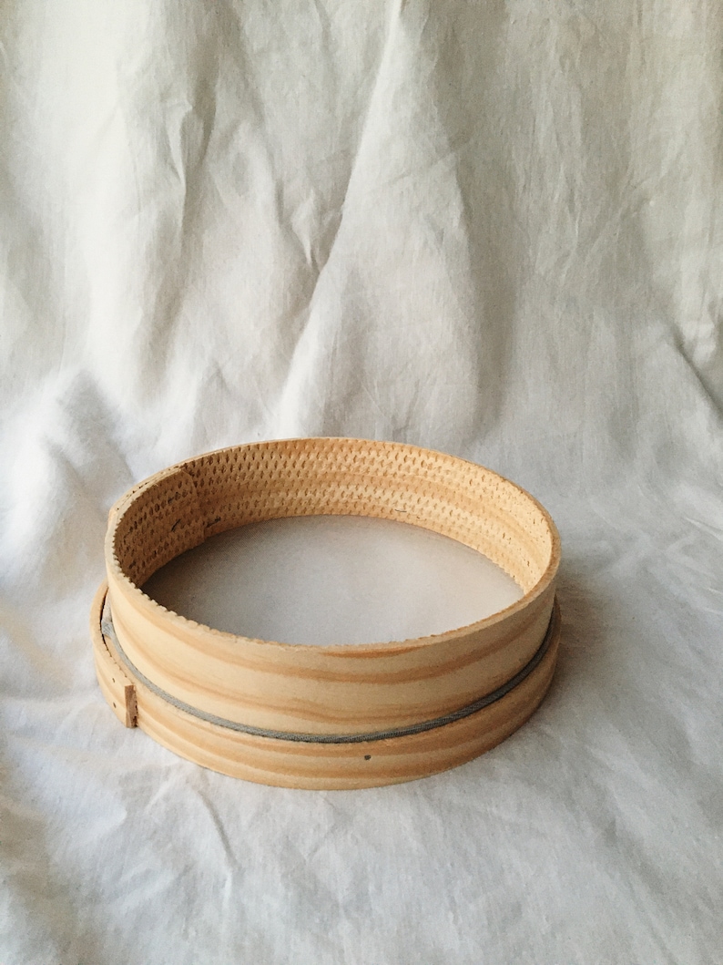 May include: A round wooden flour sifter with a gray mesh lining. The sifter is sitting on a white fabric surface.