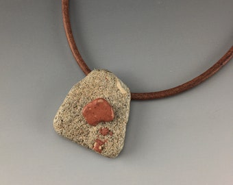 May include: A handcrafted necklace featuring a textured, light gray stone pendant with reddish-brown accents. The pendant is suspended from a brown leather cord. The necklace is a unique piece of jewelry.