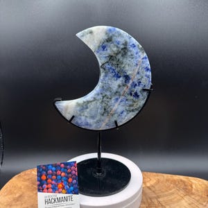 May include: A crescent moon-shaped hackmanite stone with blue, white, and grey mottled patterns, mounted on a black metal stand. A rectangular card is present with the text UV REACTIVE HACKMANITE, CROWN & THIRD EYE CHAKRA, and additional text.