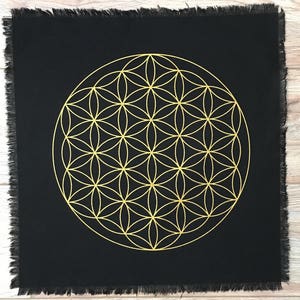 May include: A black fabric square with a gold-colored Flower of Life design. The fabric has a frayed edge.