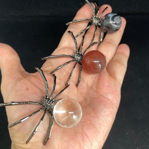 May include: Three metal spider sculptures with different colored gemstone spheres. The spiders are black metal with eight legs and a gemstone sphere in the center. The spheres are clear quartz, red carnelian, and a gray and white agate.