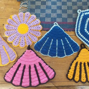 May include: Six crocheted pot holders in various colors and patterns. The pot holders are shaped like flowers, shells, and hexagons. They are made with a variety of yarn colors, including purple, blue, yellow, pink, and black.