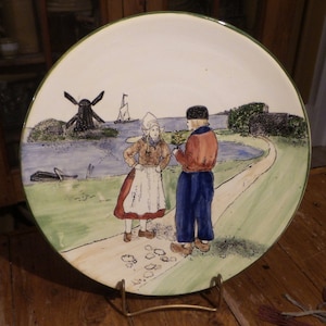 May include: A ceramic plate with a hand-painted scene of two people in traditional Dutch clothing standing on a path near a windmill and a canal. The plate has a green rim and is on a gold stand.