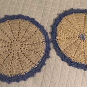 May include: Two handmade, circular crochet doilies. Each doily features a cream-colored center with a radial design and a blue scalloped border. The doilies are displayed on a white, quilted surface.