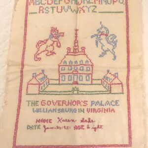 May include: Embroidered textile featuring the alphabet, a building, and two figures. The text reads "The Governor's Palace Williamsburg in Virginia" and includes a name and date. The piece is framed with a pink border.
