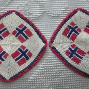 May include: Two square, crocheted trivets with a Norwegian flag design. Each trivet features a cream-colored center with four red and blue flag emblems, framed by a red and blue border. The trivets are displayed on a textured white surface.