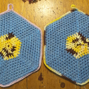 Pode incluir: Dois pegadores de panela hexagonais de crochê azul com um desenho de flor amarela, marrom e branca no centro. Um pegador de panela tem uma borda rosa e o outro tem uma borda marrom.