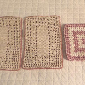 May include: Three handmade crochet items: two rectangular placemats and a square coaster. The placemats are off-white with a pink border and a central openwork design. The coaster features a pink and white geometric pattern.