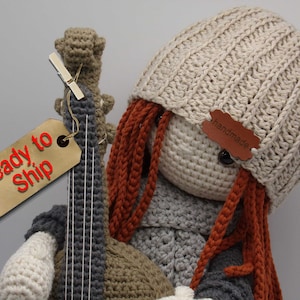 Könnte beinhalten: Häkelpuppe mit langen roten Haaren, die eine beige Strickmütze trägt und eine gehäkelte Gitarre hält. Die Puppe hat braune Augen und ein handgemachtes Etikett auf der Mütze.