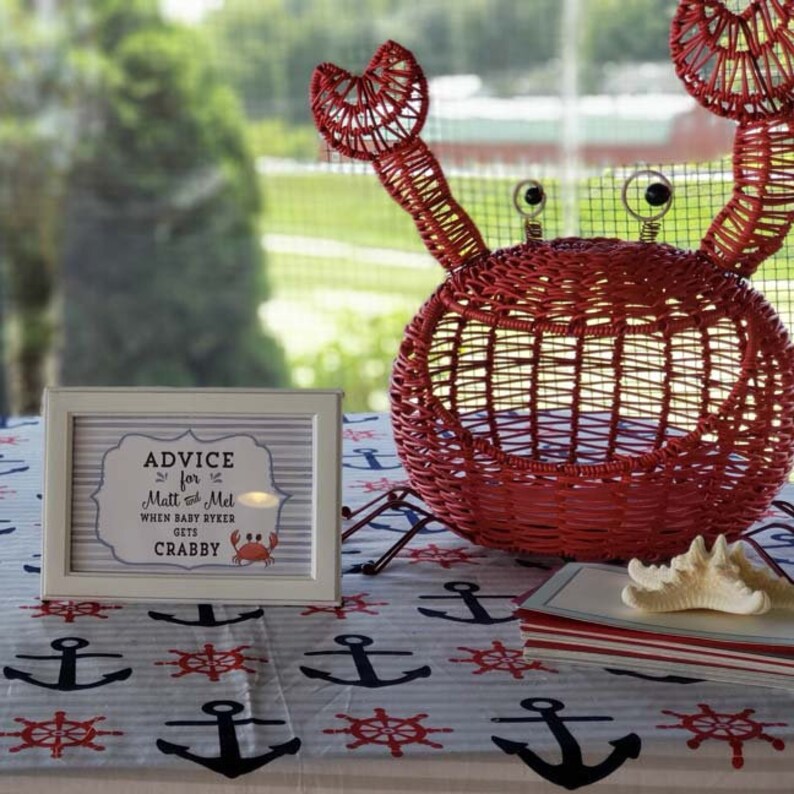 May include: A white sign with the text "Advice for Matt & Mel When Baby Ryker Gets Crabby" and a small red crab illustration. The sign is on a table with a nautical themed tablecloth and a red wicker crab.
