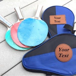 May include: A set of three blue, red, and green ping pong paddles with a black and blue carrying case. The case has two brown leather patches with the text "Your Text" on them.