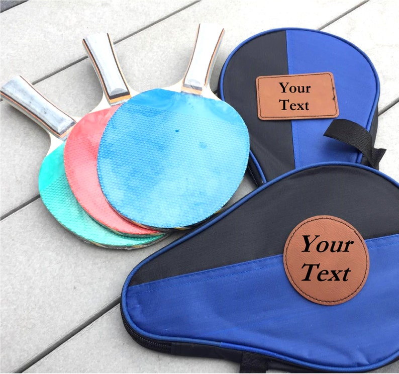 May include: A set of three blue, red, and green ping pong paddles with a black and blue carrying case. The case has two brown leather patches with the text "Your Text" on them.