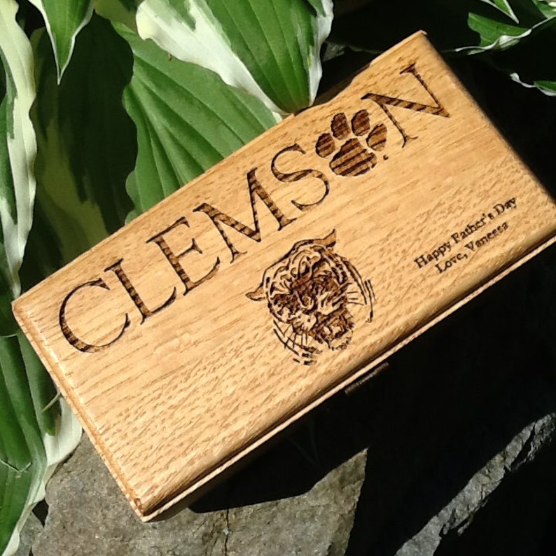 May include: A wooden box with the words 'CLEMSON' engraved on the top. The box also has a paw print and a tiger head engraved on it. The text 'Happy Father's Day Love, Vanessa' is engraved on the bottom of the box.