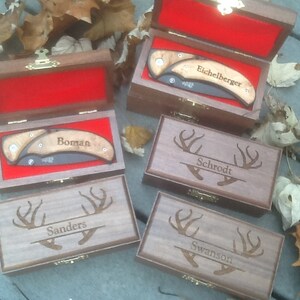 May include: Five wooden boxes with engraved deer antlers and names. Each box contains a pocket knife with a wooden handle. The names on the boxes are 'Eichelberger', 'Boman', 'Schrodt', 'Sanders', and 'Swanson'.
