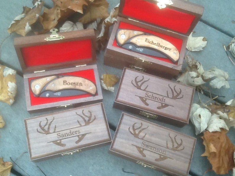 May include: Five wooden boxes with engraved deer antlers and names. Each box contains a pocket knife with a wooden handle. The names on the boxes are 'Eichelberger', 'Boman', 'Schrodt', 'Sanders', and 'Swanson'.