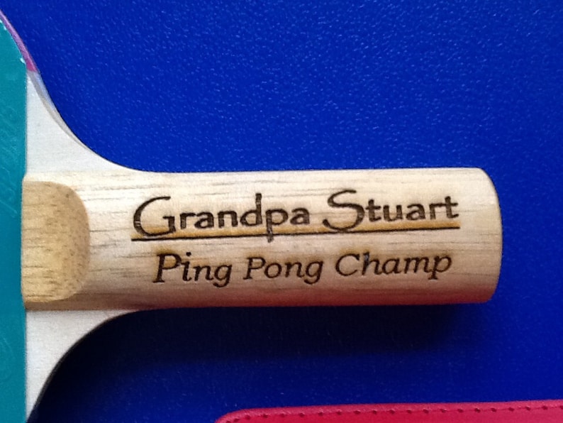 May include: A wooden ping pong paddle with a blue and green surface. The paddle is engraved with the text "Grandpa Stuart Ping Pong Champ".