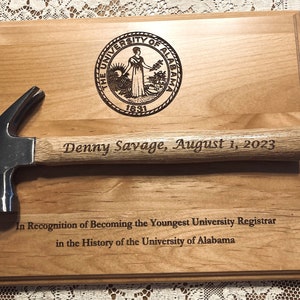 May include: A wooden plaque with a hammer engraved with "Denny Savage, August 1, 2023". The plaque also features the University of Alabama seal and the text "In Recognition of Becoming the Youngest University Registrar in the History of the University of Alabama."
