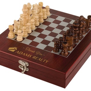 May include: A mahogany wood chess set with a hinged box. The chessboard is silver and red. The chess pieces are made of wood and are in the traditional style. The box has a gold inscription that reads "Thank You - ADAMS REALTY".