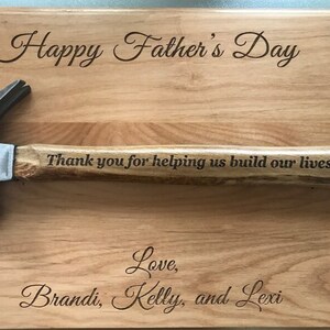 May include: A wooden cutting board with a hammer engraved with the message "Happy Father's Day. Thank you for helping us build our lives. Love, Brandi, Kelly, and Lexi."