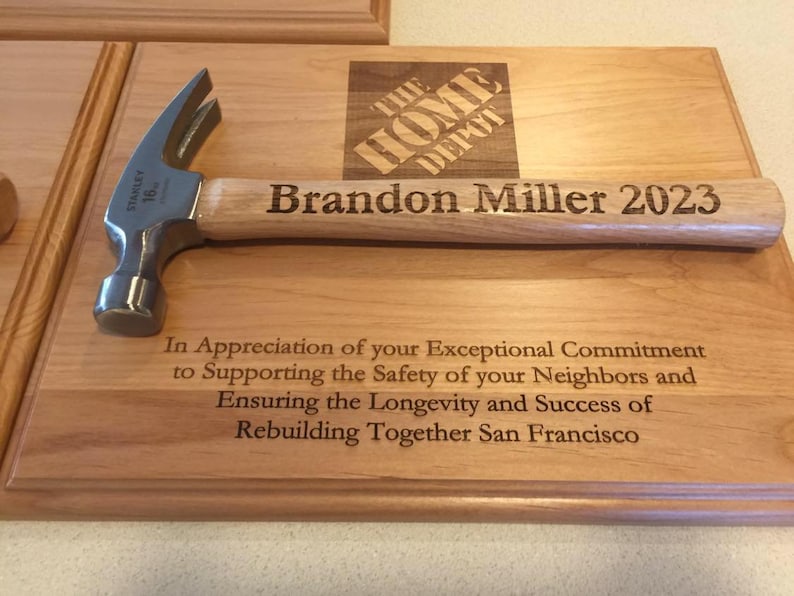 May include: A wooden plaque with a hammer on top. The plaque is engraved with the text "The Home Depot", "Brandon Miller 2023", and "In Appreciation of your Exceptional Commitment to Supporting the Safety of your Neighbors and Ensuring the Longevity and Success of Rebuilding Together San Francisco".