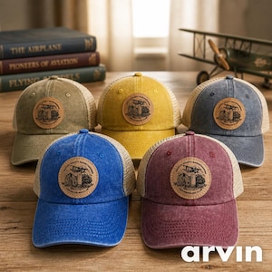 May include: Five trucker hats in various colors: olive green, yellow, blue, burgundy, and denim blue. Each hat features a circular leather patch with a graphic and text. The hats have a mesh back and a curved brim.