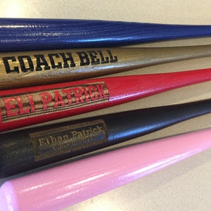 May include: Five wooden baseball bats painted in different colors, including blue, gold, red, black, and pink. Each bat has a personalized inscription, including "Coach Bell", "Eli Patrick", "Ethan Patrick Class of 2022", and "Eagles".