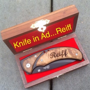 May include: A black folding knife with a wooden handle. The handle is engraved with the word 'Reiff'. The knife is in a wooden box with a red velvet lining. The box is closed and the lid is open. The lid has the text 'Knife in Ad...Reiff' in yellow letters on a red background.