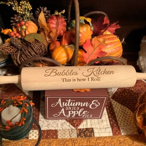May include: A wooden rolling pin with the text "Bubbles' Kitchen This is how I Roll!" engraved on it. The rolling pin is sitting on a brown and orange patterned fabric with a small wooden box that says "Autumn Skies Apple Pies" in front of it.