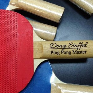 May include: A red and black ping pong paddle with a wooden handle. The handle is engraved with the text "Doug Stoffel Ping Pong Master".