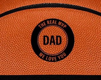 May include: An orange basketball with a textured surface and black rubber lines. A circular black emblem is printed in the center featuring the text THE REAL MVP at the top, DAD in the middle, and WE LOVE YOU at the bottom in orange lettering.