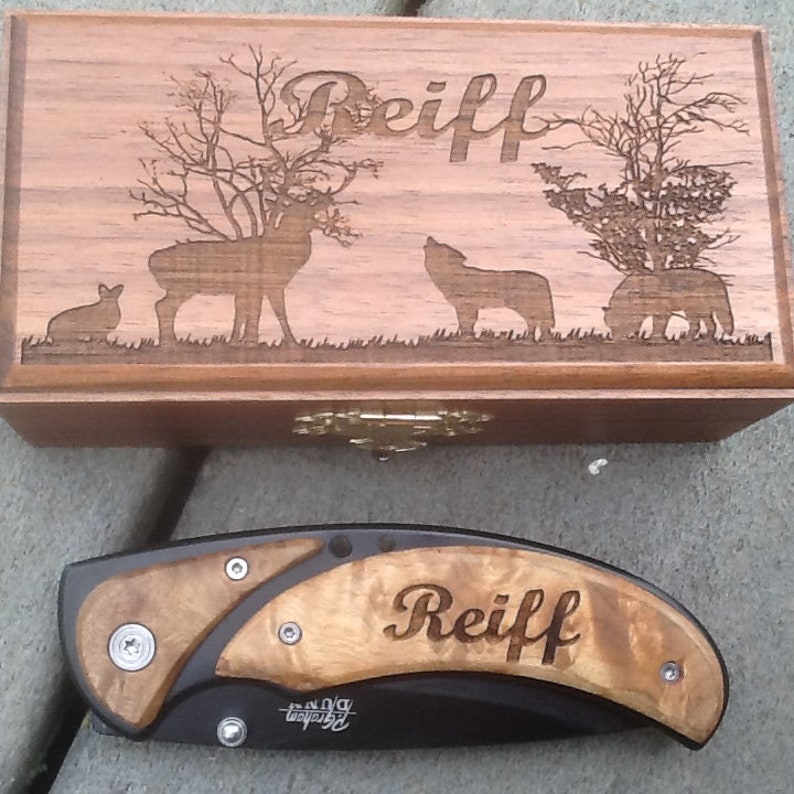 May include: A wooden box with a hinged lid and a brass latch. The box has an engraved design of a deer, a wolf, and a bear. The box is engraved with the name 'Reiff'. A black folding knife with a wooden handle is also pictured. The knife handle is engraved with the name 'Reiff'.