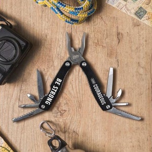 May include: Black multi-tool with a silver finish. The tool has a variety of attachments, including pliers, a knife, and a screwdriver. The tool is engraved with the words "Be Strong and Courageous".