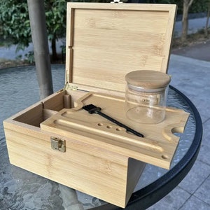May include: A light-colored bamboo box with a hinged lid, open to reveal a tray, a small glass jar with a wooden lid, and a black brush. The box has a metal clasp and hinges.