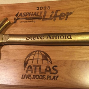 May include: A gold-colored hammer with the text "Steve Arnold" engraved on the handle. The hammer is mounted on a wooden plaque with the text "2023 Asphalt Lifer by Atlas Roofing" and "ATLAS LIVE, ROOF, PLAY".