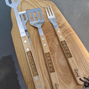 May include: A set of three grilling tools with wooden handles. The tools include a spatula, a fork, and a set of tongs. The handles are engraved with the names "Dwane" and "Boiler Up".