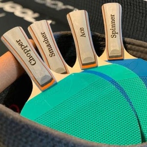 May include: Four wooden table tennis paddles with blue and green surfaces. The paddles are personalized with the names "Chopper", "Smasher", "Ace", and "Spinner".