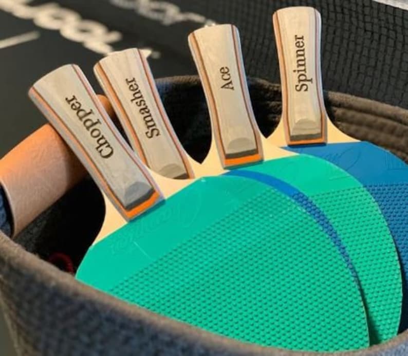May include: Four wooden table tennis paddles with blue and green surfaces. The paddles are personalized with the names "Chopper", "Smasher", "Ace", and "Spinner".