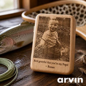May include: A wooden phone case with a laser-etched image of a baby. The case has the text "Reel grateful that you're my Pops! ~ Ronan" engraved on it. The case is on a wooden surface.
