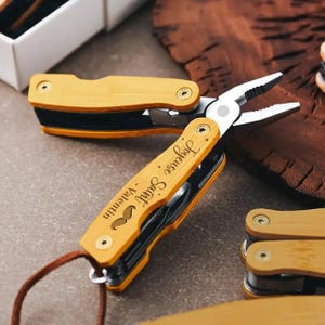 May include: A multi-tool with a light brown wooden handle and black accents. The tool includes pliers and other implements. The handle is engraved with the words "Joyeuse Saint - Valentin" and a mustache symbol. A leather cord is attached.