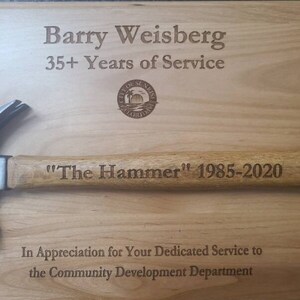 May include: A wooden plaque with the text "Barry Weisberg 35+ Years of Service" and a city seal. The plaque also has the text "The Hammer" 1985-2020 and "In Appreciation for Your Dedicated Service to the Community Development Department".