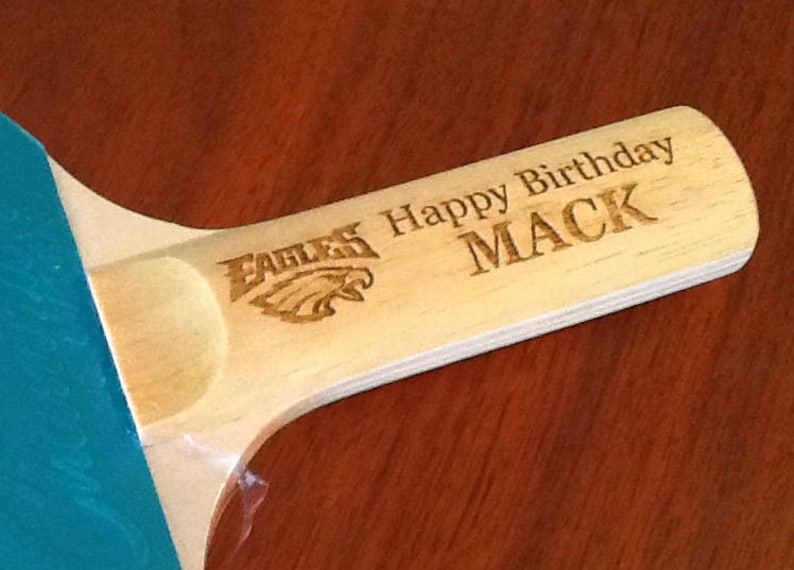 May include: A wooden table tennis paddle with a green surface and a brown handle. The handle is engraved with the words "EAGLES", "Happy Birthday", and "MACK".
