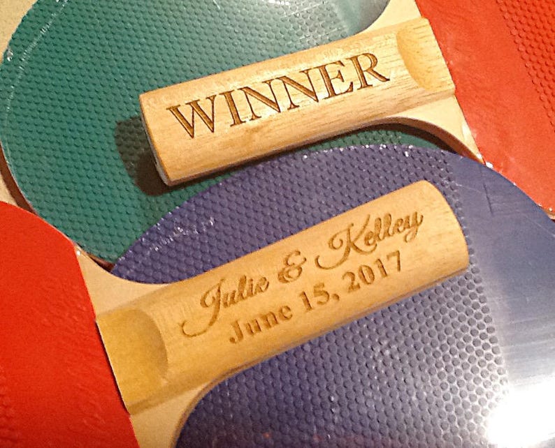 May include: Two wooden ping pong paddles with red, green, and blue surfaces. One paddle is engraved with "WINNER" and the other is engraved with "Julie & Kelley June 15, 2017".