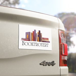 May include: White car magnet with a stack of colorful books and the text "BOOKTROVERT" in black.