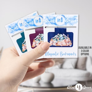 Puede incluir: Tres marcadores magnéticos con fondo azul, morado y turquesa con una ilustración de acuarela de un dragón durmiendo sobre una pila de libros. Los marcadores están etiquetados con "The Quiet Grove" y dicen "Magnetic Bookmarks" en la parte inferior. El texto "AVAILABLE IN 3 COLOR OPTIONS" está en la parte inferior derecha.