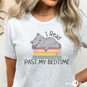 May include: Grey t-shirt featuring a grey cat napping on a pile of colourful books. The text "I Read Past My Bedtime" is printed on the shirt.