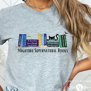 May include: A gray t-shirt with a black cat sitting on a bookshelf with a variety of books. The books have titles like "The Queen's Crown", "Hunted", "Fated", "The King's Shadow", and "The King's Captive". The text "MAGIFORD SUPERNATURAL BOOKS" is printed below the bookshelf.