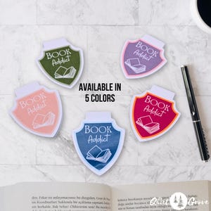 Może przedstawiać: Pięć kolorowych magnetycznych zakładek w kształcie tarczy z tekstem "BOOK Addict" i stosem książek w środku. Zakładki są dostępne w kolorze zielonym, fioletowym, różowym, czerwonym i niebieskim.