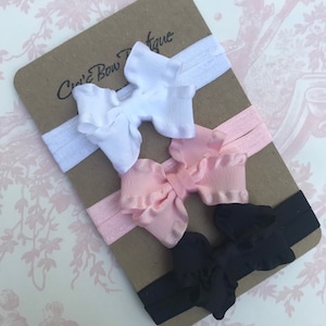 May include: Three headbands with bows in white, pink, and black. The headbands are made of stretchy elastic material. The bows are made of fabric and have a ruffled edge.
